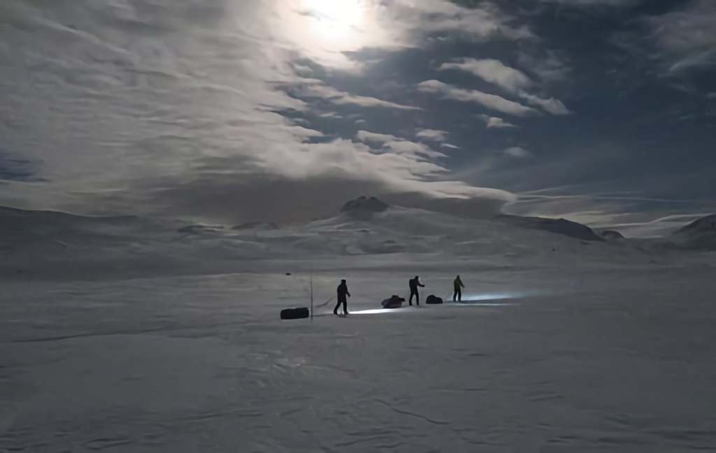 Silhouetted explorers crossing a snowy landscape under a cloudy, moonlit sky.